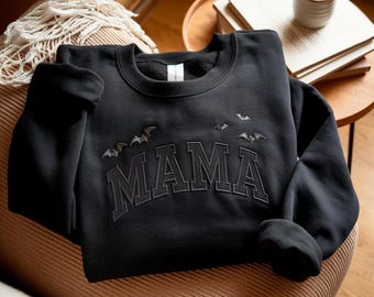 May include: Black sweatshirt with the word "MAMA" embroidered in a collegiate style font. Above the word are embroidered bats. The sweatshirt is folded on a brown surface. A cozy, fall-themed garment.