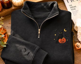 Könnte beinhalten: Ein schwarzes Sweatshirt mit kurzem Reißverschluss und einer Stickerei, die eine schwarze Katze neben einem orangefarbenen Kürbis zeigt, ergänzt durch einen Halbmond, Sterne und kleine Fledermäuse. Weitere Fledermäuse sind am Ärmel gestickt.
