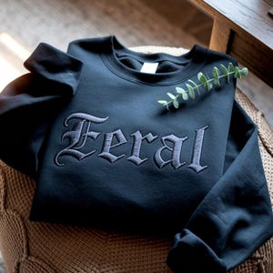 May include: A black crewneck sweatshirt with the word "Feral" embroidered in a gothic font across the chest. The sweatshirt is folded and resting on a woven surface, with a sprig of greenery placed on top.
