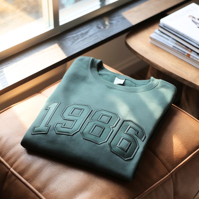 K&ouml;nnte beinhalten: Ein salbeigr&uuml;nes Sweatshirt mit Rundhalsausschnitt und der Zahl 1986 in gro&szlig;en, dunkelgr&uuml;n gestickten Blockbuchstaben auf der Brust. Das Kleidungsst&uuml;ck hat einen gerippten Kragen und lange &Auml;rmel, wobei das weiche Fleece-Innenfutter am Halsausschnitt sichtbar ist.