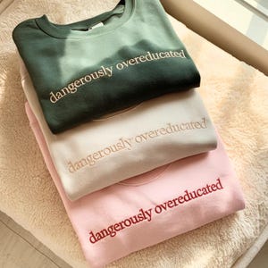 May include: Three folded sweatshirts in shades of green, cream, and pink are stacked on a soft, textured surface. Each sweatshirt has the embroidered text "dangerously overeducated" in a contrasting color. The sweatshirts are neatly arranged, showcasing the text.