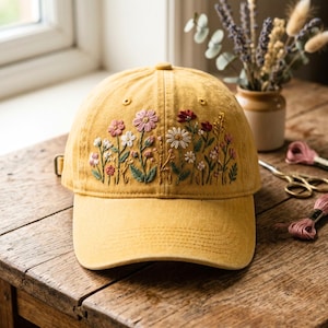 May include: Mustard yellow baseball cap with embroidered floral design. The embroidery features a variety of flowers in pink, white, and red, with green leaves. The cap has a curved brim and a metal clasp closure.