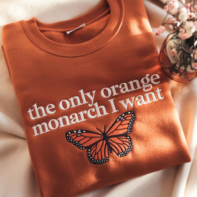 May include: An orange sweatshirt with the text "the only orange monarch I want" in white. A detailed embroidered monarch butterfly is below the text. The sweatshirt is folded on a cream-colored surface.
