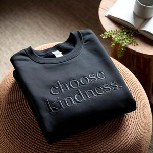 May include: A black sweatshirt with the embroidered phrase "choose kindness." The crew neck sweatshirt is folded and resting on a woven stool. The text is in a simple, elegant font.