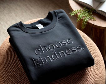 May include: A black sweatshirt with the embroidered phrase "choose kindness." The crew neck sweatshirt is folded and resting on a woven stool. The text is in a simple, elegant font.