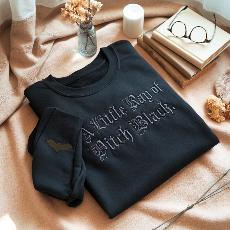 May include: Black sweatshirt and matching pants set. The sweatshirt features the embroidered text "A Little Ray of Pitch Black." The pants have a bat graphic. The set is folded on a peach-colored blanket.