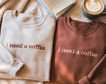 May include: Two crewneck sweatshirts, one beige and one rust-colored, embroidered with the phrase "I need a coffee" in a complementary color. A cup of coffee with latte art and an open book are in the background.
