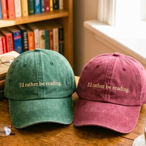 May include: Two baseball caps, one in forest green and one in maroon, both featuring a distressed fabric texture. Each cap has the text "I'd rather be reading." embroidered in a light cream colour across the front panel above the curved brim.