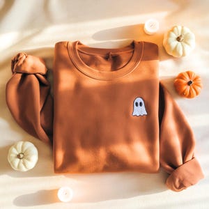 Puede incluir: Una sudadera de color óxido con un parche de fantasma blanco. La imagen incluye pequeñas calabazas y velas, lo que sugiere un tema otoñal o de Halloween.