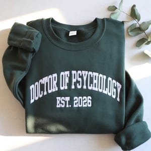 May include: A dark green sweatshirt with the text "DOCTOR OF PSYCHOLOGY EST. 2026" in white, arched across the chest. The sweatshirt has a crew neck and long sleeves. The sleeves are cuffed. The background is a light gray.
