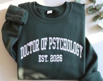 May include: A dark green sweatshirt featuring the white text "DOCTOR OF PSYCHOLOGY EST. 2026". The text is arched across the chest. The sweatshirt has a crew neck and long sleeves with cuffed ends. The background is a light grey.