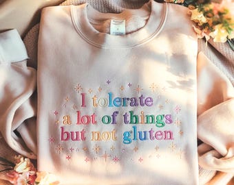 Puede incluir: Sudadera rosa claro con la frase bordada "I tolerate a lot of things but not gluten" en colores arcoíris. El diseño incluye pequeños acentos de estrellas y cruces. La sudadera se muestra sobre una superficie de tono neutro.