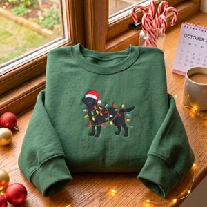 May include: A forest green crewneck sweatshirt featuring an embroidered black Labrador retriever wearing a red and white Santa hat, wrapped in colorful Christmas string lights. The design is centered on the chest of the long-sleeved garment.