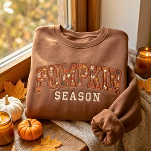 May include: A brown crewneck sweatshirt featuring the word PUMPKIN embroidered in a block font filled with a colorful floral and leaf pattern, with the word SEASON embroidered below in a simple, solid cream-colored sans-serif typeface.