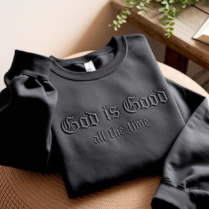 May include: A dark gray sweatshirt with the embroidered text "God is Good all the time" in a gothic font. The sweatshirt is folded, with the sleeves visible. The fabric appears soft and comfortable.