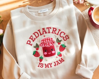May include: Cream-colored sweatshirt with the embroidered text "PEDIATRICS IS MY JAM" in red. The design features a smiling jar of jam surrounded by strawberries and leaves. The sweatshirt has a crew neck and long sleeves.