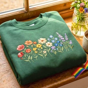 May include: A folded, forest green sweatshirt with a crew neck. The front features an embroidered floral design with various colorful flowers, including orange, yellow, white, blue, and purple. The sweatshirt is folded neatly on a wooden surface.
