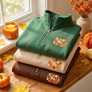 May include: A stack of three quarter-zip sweatshirts in forest green, cream, and dark brown. Each sweatshirt features an embroidered pumpkin design on the chest, composed of small orange, yellow, and white flowers with green stems.