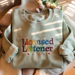 May include: A light green sweatshirt with the text "Licensed Listener" embroidered in rainbow colors. The text is surrounded by small stars and hearts.