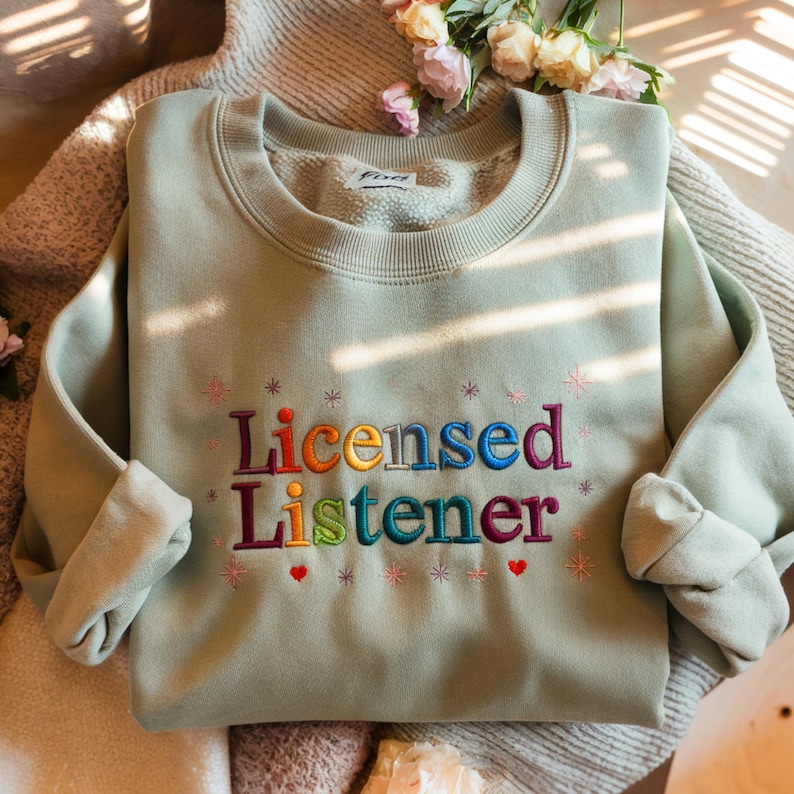 May include: A light green sweatshirt with the text "Licensed Listener" embroidered in rainbow colors. The text is surrounded by small stars and hearts.
