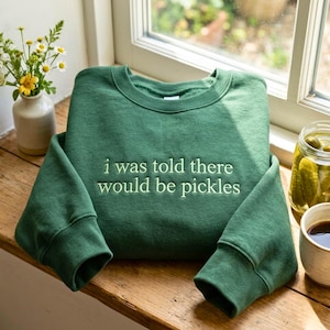 Puede incluir: Una sudadera de cuello redondo en color verde bosque con el texto bordado "i was told there would be pickles" en hilo verde claro. La prenda tiene mangas largas, cuello acanalado y un corte clásico, sobre una superficie de madera.