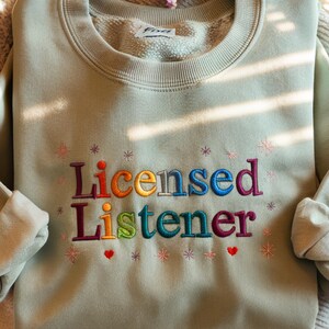 May include: A light green sweatshirt with the words "Licensed Listener" embroidered in rainbow colors. The text is surrounded by small white stars and two red hearts.