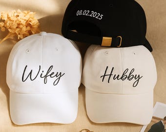 Może przedstawiać: Trzy czapki z daszkiem: czarna z napisem "08.02.2025", biała z napisem "Wifey" i kremowa z napisem "Hubby" wyszytym czarną czcionką. Czapki mają regulowane paski i złotą klamrę. Widoczne są dwa złote pierścionki.