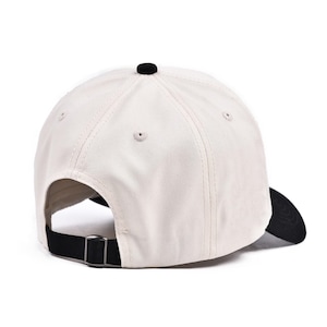 May include: A cream-colored baseball cap with a black brim and button. The adjustable strap is black with a silver-colored metal buckle. The cap is made of a sturdy fabric.