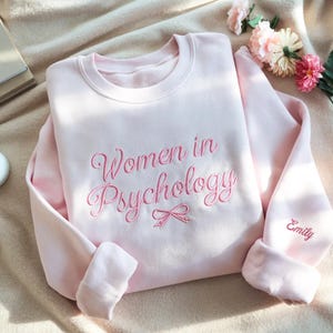 May include: A light pink sweatshirt with the embroidered text "Women in Psychology" in a cursive font. A small bow is embroidered below the text. The name "Emily" is embroidered on the sleeve. The sweatshirt is folded on a beige surface.