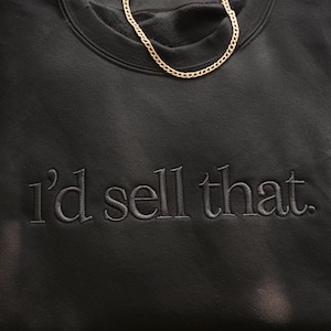 K&ouml;nnte beinhalten: Schwarzes Sweatshirt mit dem gestickten Satz "I'd sell that." Eine goldene Halskette liegt auf der Oberseite des Sweatshirts. Das Sweatshirt hat einen Rundhalsausschnitt und ein weiches, strukturiertes Aussehen.