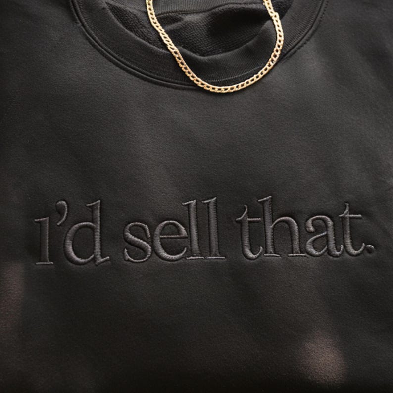 K&ouml;nnte beinhalten: Schwarzes Sweatshirt mit dem gestickten Satz "I'd sell that." Eine goldene Halskette liegt auf der Oberseite des Sweatshirts. Das Sweatshirt hat einen Rundhalsausschnitt und ein weiches, strukturiertes Aussehen.