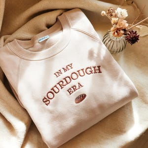 May include: A light pink sweatshirt with the embroidered phrase "IN MY SOURDOUGH ERA" in a brown color. A small embroidered bread loaf is below the text. The sweatshirt is folded on a beige blanket.