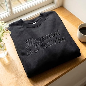 Puede incluir: Una sudadera negra con la frase "Movement is Medicine" bordada en un tono a juego. La sudadera de cuello redondo está doblada sobre una superficie de madera cerca de una ventana. También se ve una taza de café.