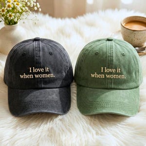 May include: Two baseball caps, one charcoal gray and one olive green, both featuring a six-panel design with a curved brim and an adjustable strap. Each cap has the text "I love it when women." embroidered in a light beige thread on the front.