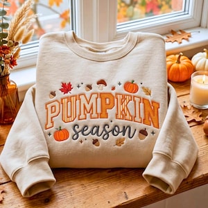 May include: A beige crewneck sweatshirt featuring embroidered text that reads PUMPKIN season in orange and dark grey. The design is decorated with embroidered autumn motifs including orange pumpkins, brown acorns, red maple leaves, and small mushrooms.