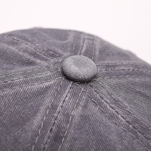 May include: Close-up of a gray, textured newsboy cap with a circular button on top. The cap features visible stitching and a classic design, suitable for various styles. The fabric appears to be a soft, durable material.