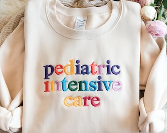 May include: Cream-colored sweatshirt with the embroidered text "pediatric intensive care" in rainbow colors. The crew neck sweatshirt is made of a soft material and has long sleeves. The design is centered on the front of the garment.