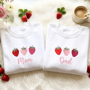 May include: Two white crewneck sweatshirts featuring embroidered strawberry designs. The left sweatshirt has three strawberries and the text Mom in pink thread. The right sweatshirt has three strawberries and the text Dad in pink thread.