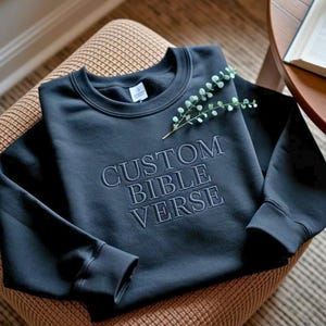 May include: A navy blue sweatshirt with the embroidered text "CUSTOM BIBLE VERSE." The sweatshirt is displayed on a patterned chair and adorned with a sprig of greenery. The design features a crew neck and ribbed cuffs.