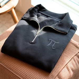 Könnte beinhalten: Ein schwarzes Sweatshirt mit silbernem Reißverschluss und gesticktem Pi-Symbol. Das Sweatshirt liegt gefaltet auf einem braunen Kissen. Im Hintergrund ist ein Holzstuhl zu sehen.