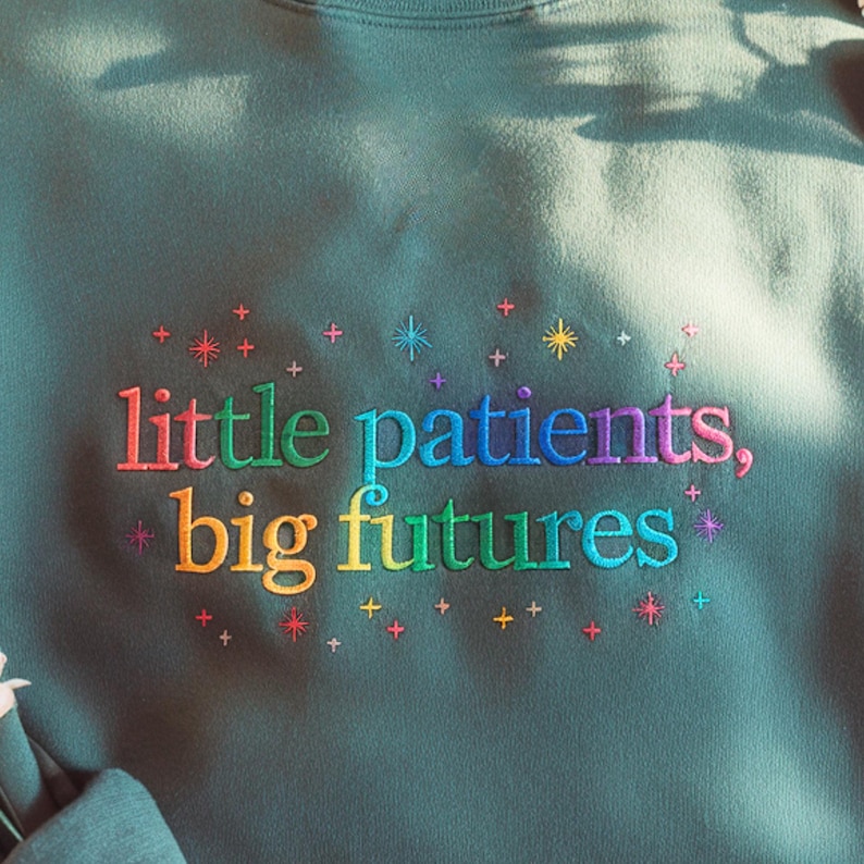 Puede incluir: Sudadera de color verde azulado con texto bordado en colores arco&iacute;ris: "little patients, big futures." El texto est&aacute; rodeado de peque&ntilde;as estrellas y cruces bordadas. Una prenda superior informal y c&oacute;moda.