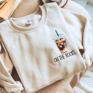 May include: Cream-colored sweatshirt featuring an embroidered iced coffee design with a green straw. The phrase "ON THE ROCKS" is embroidered below the coffee cup in black. The sweatshirt is folded on a neutral-toned surface.