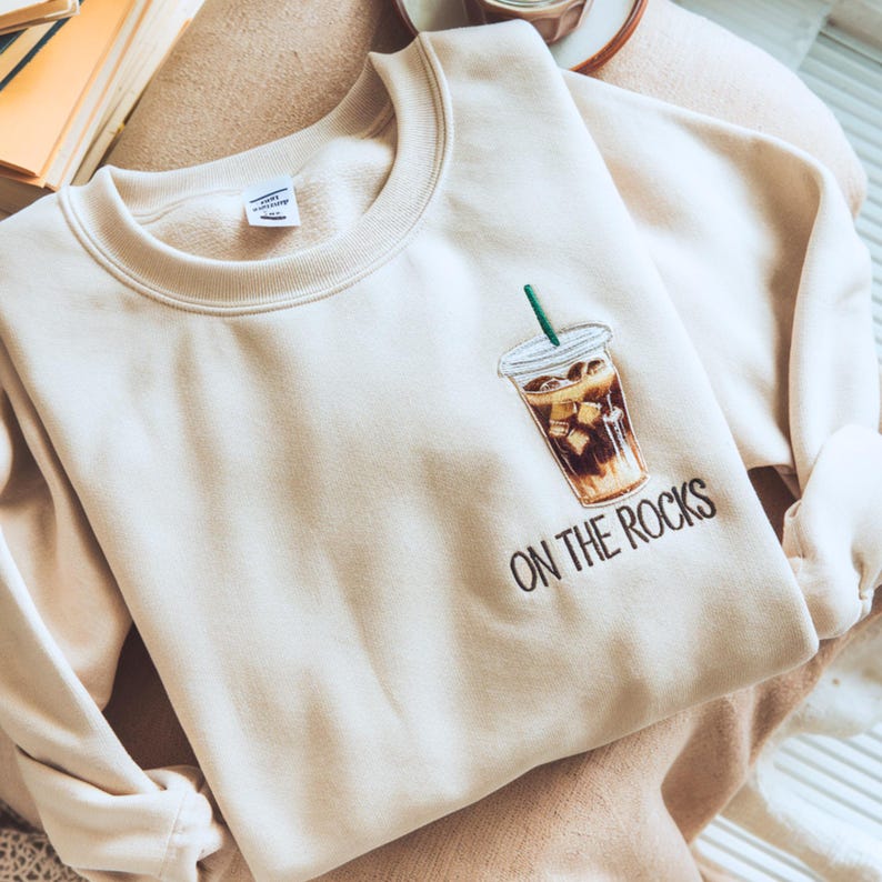 May include: Cream-colored sweatshirt featuring an embroidered iced coffee design with a green straw. The phrase "ON THE ROCKS" is embroidered below the coffee cup in black. The sweatshirt is folded on a neutral-toned surface.
