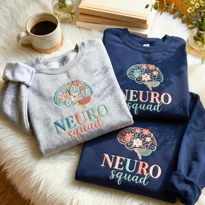 May include: Three sweatshirts are shown, two navy and one grey. Each has an embroidered brain design with flowers and the text "NEURO squad". A coffee mug and flowers are in the background.