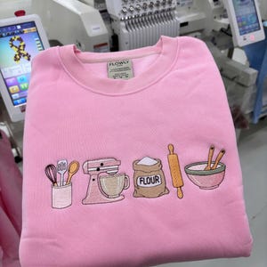 May include: A light pink crewneck sweatshirt featuring an embroidered design of baking tools including a utensil holder, a stand mixer, a sack labeled FLOUR, a rolling pin, and a mixing bowl with spoons. The interior neck label reads FLOWLY.