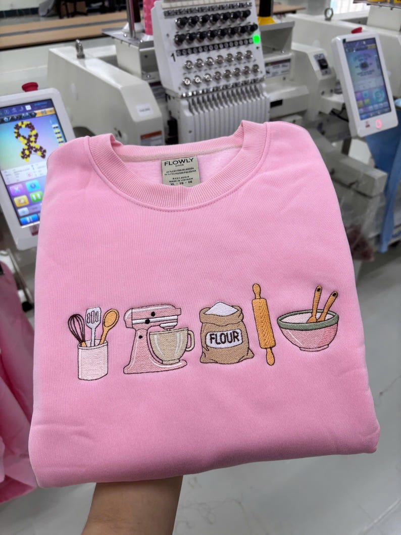 May include: A light pink crewneck sweatshirt featuring an embroidered design of baking tools including a utensil holder, a stand mixer, a sack labeled FLOUR, a rolling pin, and a mixing bowl with spoons. The interior neck label reads FLOWLY.
