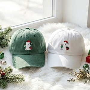 May include: Two baseball caps, one in forest green and one in white, each featuring an embroidered design of a white ghost wearing a red Santa hat, holding a green coffee cup in one hand and a red and green wrapped gift in the other hand.