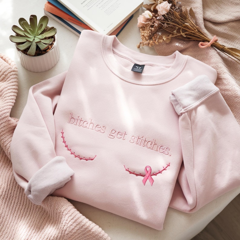 Pode incluir: Uma camisola de gola redonda em rosa claro com o texto bordado "bitches get stitches." em fio rosa a condizer. Abaixo do texto, existem duas linhas curvas de pontos de sutura bordados em rosa, com um pequeno la&ccedil;o rosa &agrave; direita.