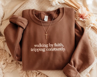 May include: A brown sweatshirt with the embroidered text "walking by faith, tripping constantly." A gold-colored cross necklace rests on the sweatshirt. The sweatshirt is folded on a cream-colored blanket, with dried flowers in the background.