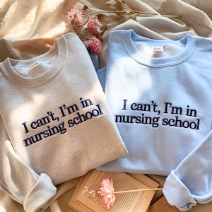 May include: Two crewneck sweatshirts, one light gray and one light blue, embroidered with the text "I can't, I'm in nursing school" in navy blue thread. The sweatshirts are displayed on a beige fabric with a book and pink flowers.